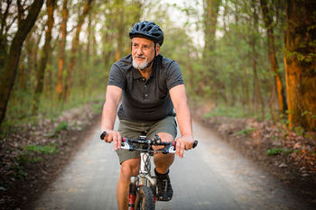 Ein vitaler Mann in höherem Alter fährt Fahrrad auf einem Waldweg. Er trägt Sportsachen und einen Helm, hat einen grauen Bart und blickt in die Kamera.