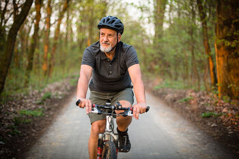 Ein vitaler Mann in höherem Alter fährt Fahrrad auf einem Waldweg. Er trägt Sportsachen und einen Helm, hat einen grauen Bart und blickt in die Kamera.