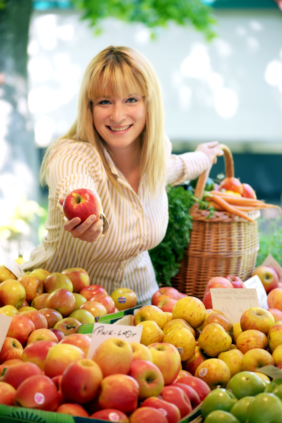 Eine freundliche Frau steht hinter einem Marktstand mit verschiedenen Apfelsorten und hält dem Betrachter einen Apfel hin. Hinter ihr steht ein geflochtener Korb, aus dem Karotten und Äpfel herausgucken.