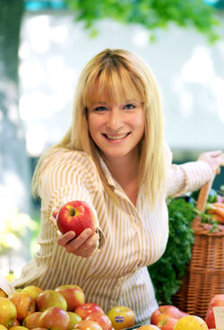 Eine freundliche Frau steht hinter einem Marktstand mit verschiedenen Apfelsorten und hält dem Betrachter einen Apfel hin. Hinter ihr steht ein geflochtener Korb, aus dem Karotten und Äpfel herausgucken.