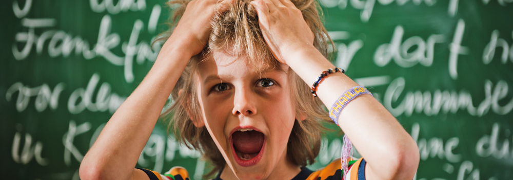 Ein ungefähr elfjähriger Junge in einem bunten T-Shirt steht vor einer beschriebenen grünen Schultafel. Er greift mit beiden Händen in seine mittellangen lockigen Haare und hat scherzhaft-erschrocken den Mund geöffnet. Er benötigt einen Notfallplan für Kopfläuse. 
