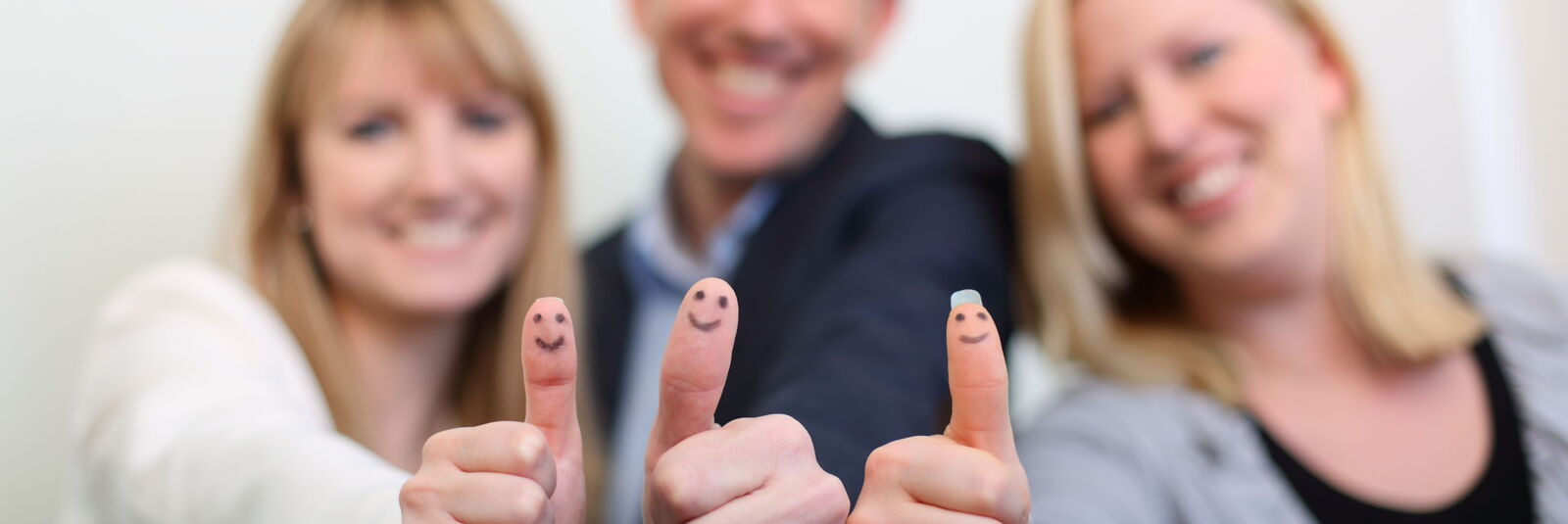 Zwei Frauen und ein Mann stehen nebeneinander und zeigen dem Betrachter jeweils die "Daumen hoch"-Geste und lächeln. Auf die Fingerkuppen ihrer Daumen ist jeweils ein Smiley gemalt. 