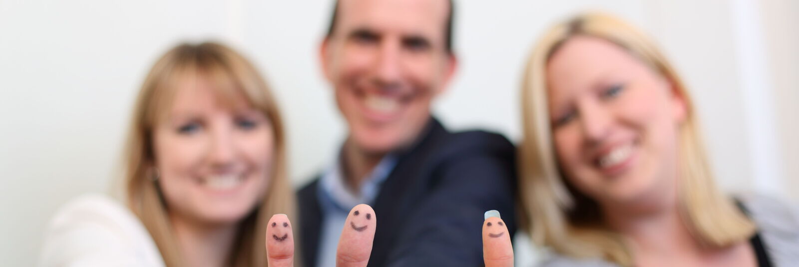 Zwei Frauen und ein Mann stehen nebeneinander und zeigen dem Betrachter jeweils die "Daumen hoch"-Geste und lächeln. Auf die Fingerkuppen ihrer Daumen ist jeweils ein Smiley gemalt. 