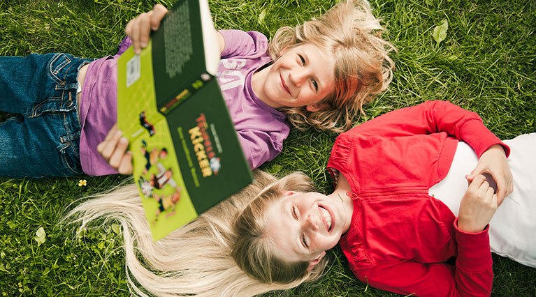 Zwei ungefähr elfjährige Kinder, ein Junge und ein Mädchen, liegen nebeneinander auf einer Wiese und lachen. Der Junge hält ein Buch in die Höhe.  