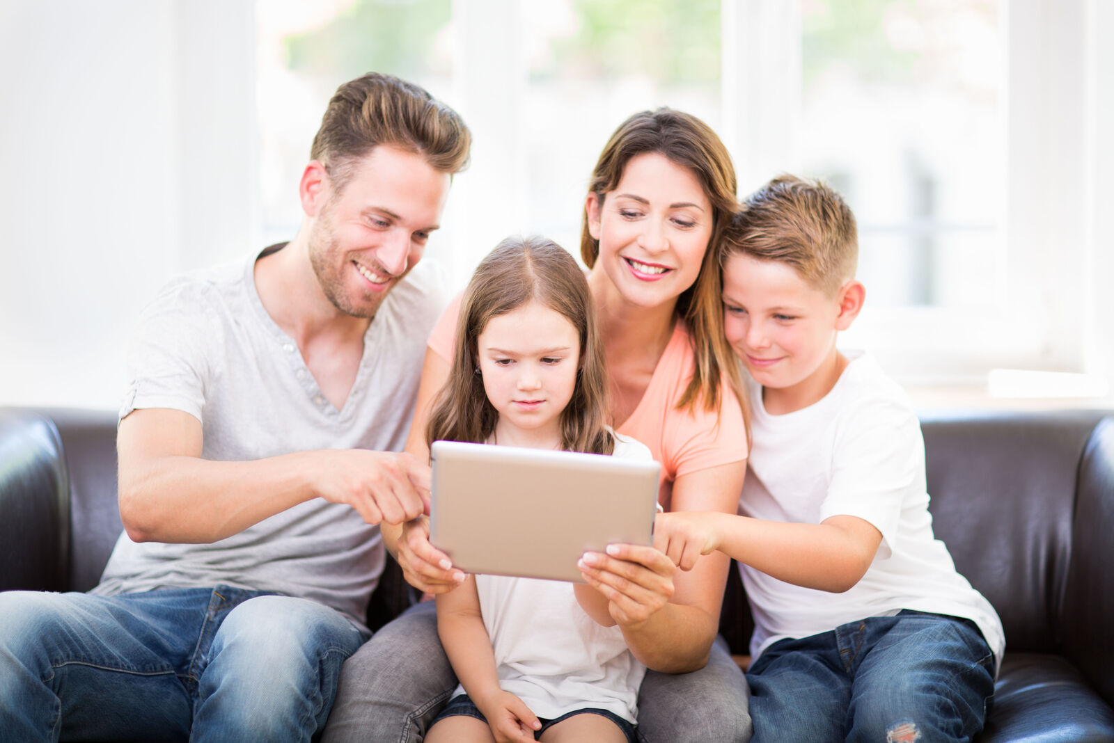 Eine fröhliche Familie mit zwei Kindern sitzt gemeinsam auf einem Ledersofa im Wohnzimmer und schaut gemeinsam auf ein Tablet.
